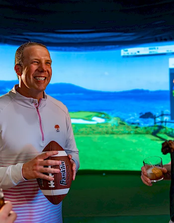 A group of people are socializing indoors, holding drinks, with one person holding a football in front of a golf simulator screen.
